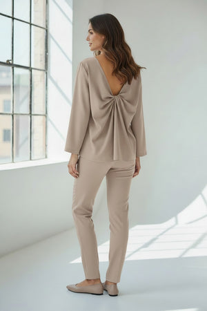 A person wears the MEIMEIJ TOP WITH KNOT BACK DETAIL in bright orange, paired with matching pants, standing against a plain light grey background. Their face is partially out of frame.
