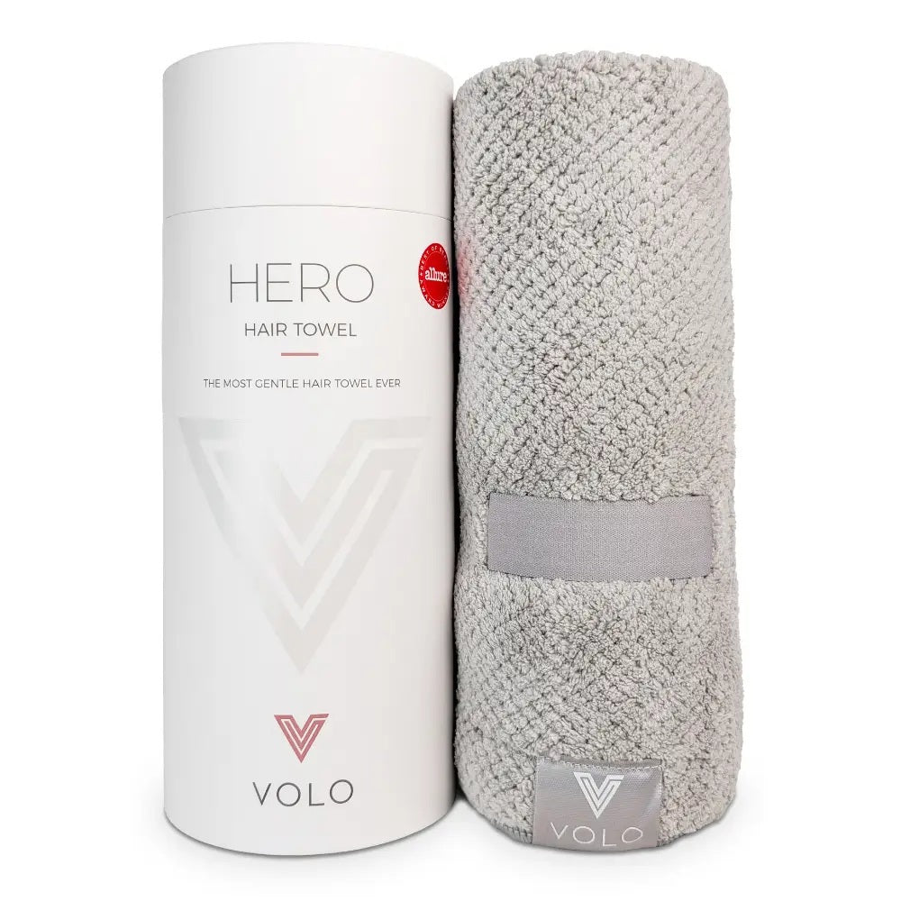 A person with short dark hair, wearing a white shirt, wraps their hair in the VOLO BEAUTY - HERO HAIR TOWEL LUXE and adjusts it with both hands in a bright, airy space filled with soft natural light.