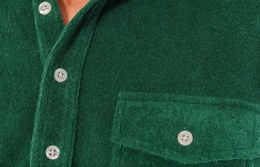 A smiling man with wavy brown hair wears the CRIQUET Terrycloth Players Shirt in Alligator Alley, featuring buttons and a chest pocket, paired with light-colored pants, standing against a white background.
