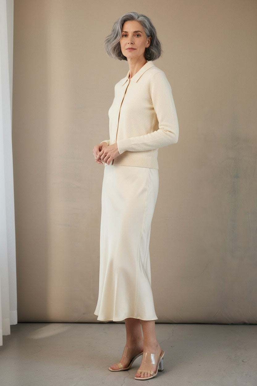 A woman with wavy brown hair wears a black short-sleeve V-neck top, black heels, and the ROSE & CROWN ROSE AND CROWN Satin Midi Skirt, standing and smiling against a plain white background.