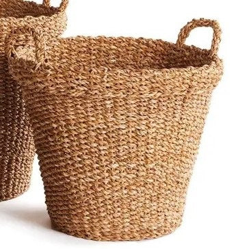 A NAPA HOME AND GARDEN Seagrass Tapered Basket with Handles and Cuff (Medium) sits on a woven rug, holding tall decorative green grass, with a chair and colorful painting in the background.