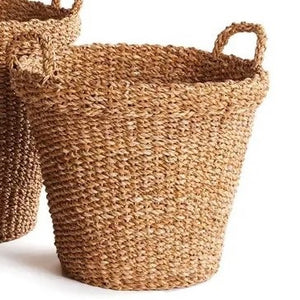 A NAPA HOME AND GARDEN Seagrass Tapered Basket with Handles and Cuff (Medium) sits on a woven rug, holding tall decorative green grass, with a chair and colorful painting in the background.