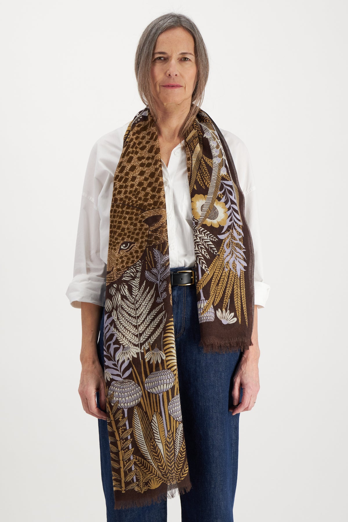 A person with shoulder-length gray hair stands facing the camera, wearing a white shirt and blue jeans. Adorning their neck is an INOUI EDITIONS - ROUSSEAU WOOL SCARF by INOUI, featuring an artisan-made brown fabric with a floral and animal pattern. The background is plain white.