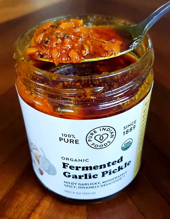 An open jar of PURE INDIAN FOODS-FERMENTED GARLIC PICKLE with a spoonful of chunky, reddish-orange fermented garlic held above it. The label notes it&#39;s mildly garlicky and moderately spicy. Brand: PURE INDIAN FOODS.
