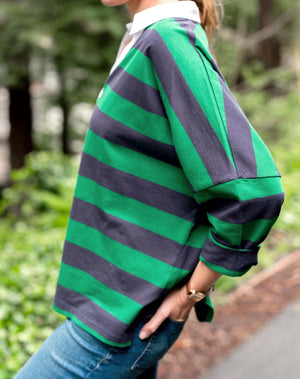 A person wears the MERSEA Amelia Rugby Tee in navy green stripe, featuring a white collar, and blue jeans. They stand outdoors with hands in pockets, green foliage visible behind them.