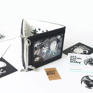 A TUMMY TIME GALLERY soft book from WEE GALLERY, featuring black and white fish and squid illustrations, is partially inserted in a clear front pocket. A brown tag attached by a string reads "tummy time gallery." This Montessori toy, ideal for sensory development, rests on a plain white surface.