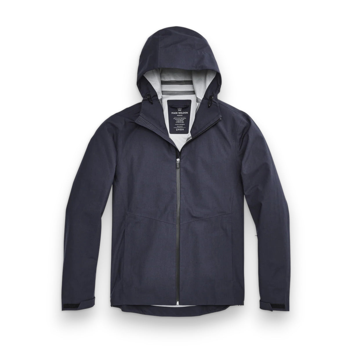 A man stands in front of a white wall wearing the MACK WELDON STORMCHASER JACKET with adjustable hood, paired with a gray shirt, blue pants, and black sneakers. He looks to the side while holding the jacket's zipper.