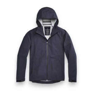 A man stands in front of a white wall wearing the MACK WELDON STORMCHASER JACKET with adjustable hood, paired with a gray shirt, blue pants, and black sneakers. He looks to the side while holding the jacket's zipper.