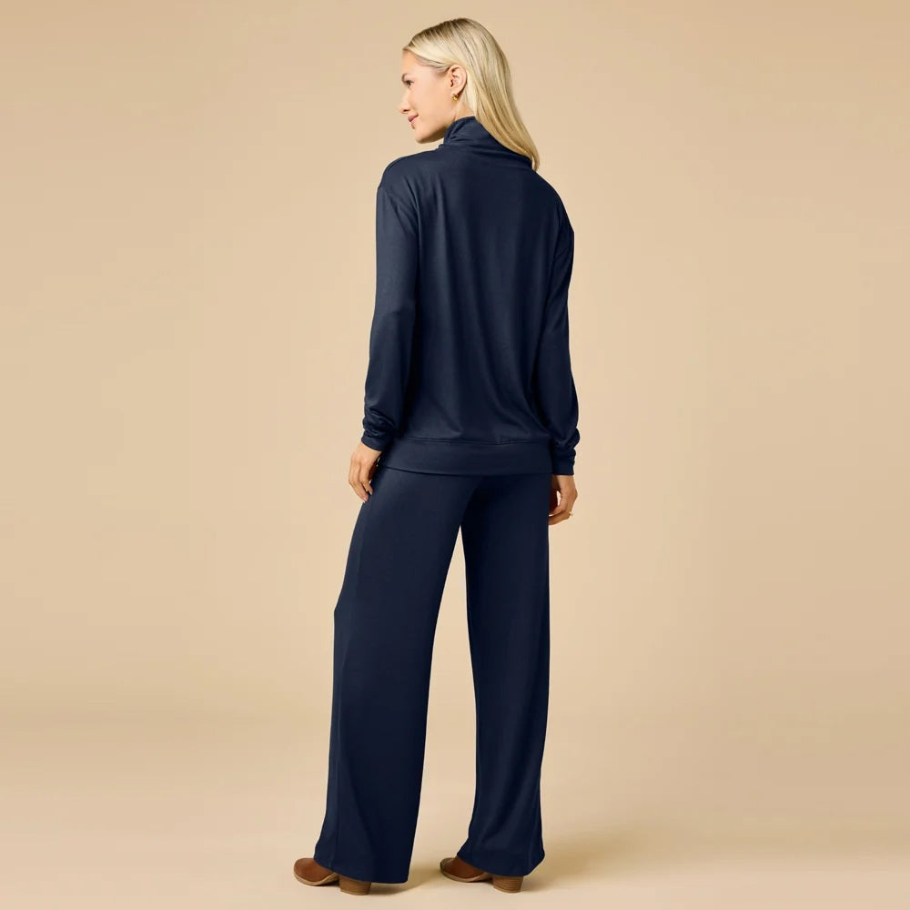 A woman wears a navy blue SOFTIES - DREAM JERSEY COWL NECK LOUNGE SET consisting of a long-sleeved top and wide-leg trousers.
