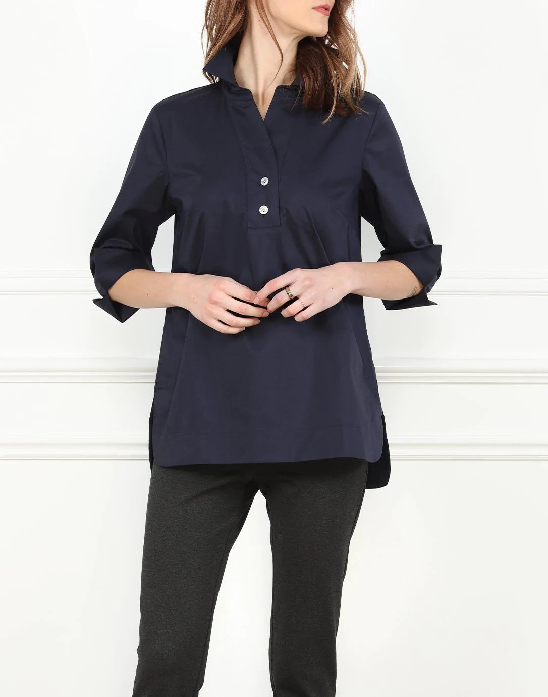 Sporting long hair, the individual is clad in a white HINSON WU - AILEEN TUNIC and black pants. The tunic-length shirt by Hinson Wu features a collar and two buttons near the top. They stand in front of a light gray wall with white molding, with their face not visible in the image.