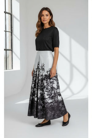 A woman wears a sheer, floral blouse paired with the MILVA MI - LONG MAXI SKIRT by MILVA—a shiny, high-waist skirt featuring a colorful floral print—and pointed black heels, posing against a simple gray studio backdrop.