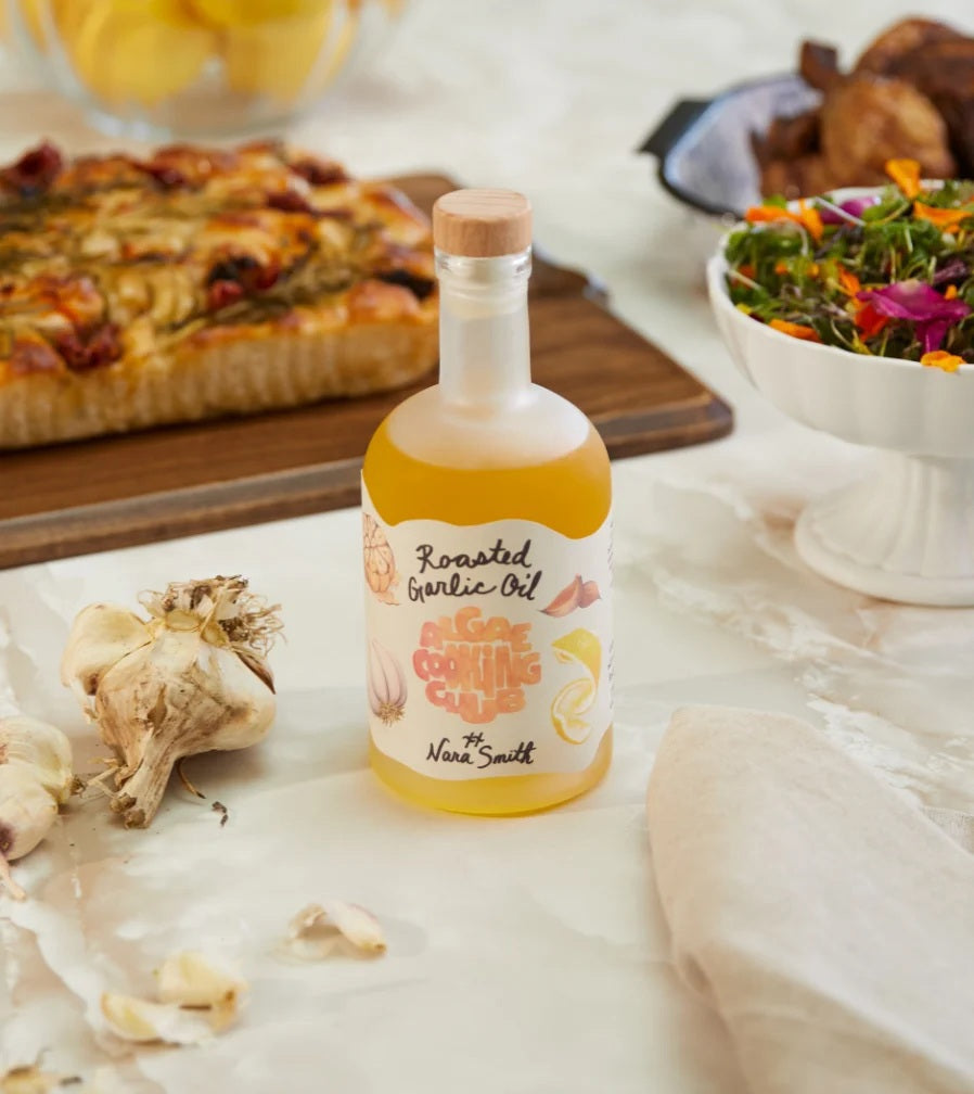 A bottle of ALGAE COOKING CLUB - NARA’S ROASTED GARLIC OIL with a tan label featuring the ALGAE COOKING CLUB logo, garlic illustrations, and “Nara Smith” signed at the bottom, stands against a white background. This rich blend uses premium algae oil for a uniquely flavorful and innovative culinary experience.