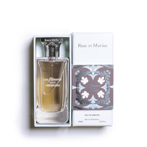 A clear glass ROSE ET MARIUS bottle, labeled "A STROLL UNDER THE ORATORY EAU DE PARFUM 100 ML," features a silver spray top, square cap, and a "Unisexe" label—capturing the floral essence of this unisex fragrance.