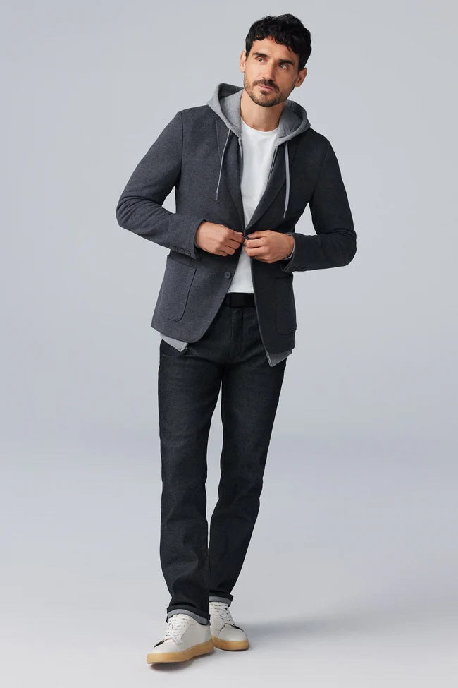 A man with short dark hair and a trimmed beard wears the MACK WELDON Ace Blazer in Total Eclipse over a white t-shirt, looking slightly to the side while buttoning his blazer against a plain light background.