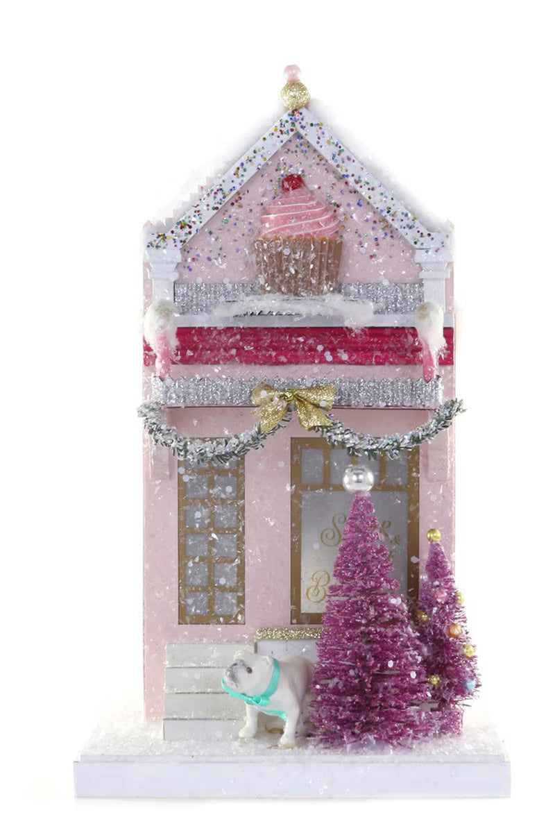 The SUGAR AND SPICE BAKE HOUSE by CODY FOSTER is a pink, snow-covered, 11-inch cardboard bakery with a cupcake roof decoration, two pink Christmas trees, silver garland, and a white dog in a blue scarf standing out front.