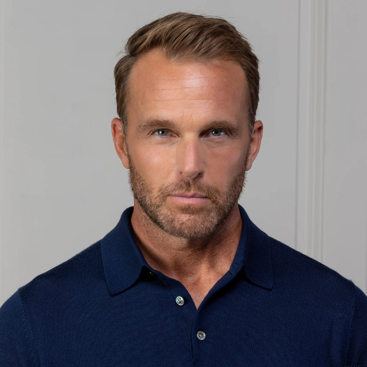 A man with short light brown hair wears the COLLARS AND CO Bellagio Three Button Long Sleeve Merino Polo in Navy and beige pants, standing before a paneled wall, holding eyeglasses and looking at the camera.