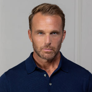 A man with short light brown hair wears the COLLARS AND CO Bellagio Three Button Long Sleeve Merino Polo in Navy and beige pants, standing before a paneled wall, holding eyeglasses and looking at the camera.