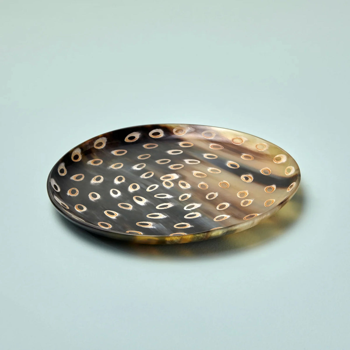 The PEACOCK HORN PLATE MEDIUM by BE HOME, INC is a round, shallow metal plate with a smooth surface, adorned with a pattern of evenly spaced, small circular indentations. Handmade in Vietnam, this plate features a slightly reflective finish with a warm goldish hue and subtle variations in color. It is set against a soft, muted green background.
