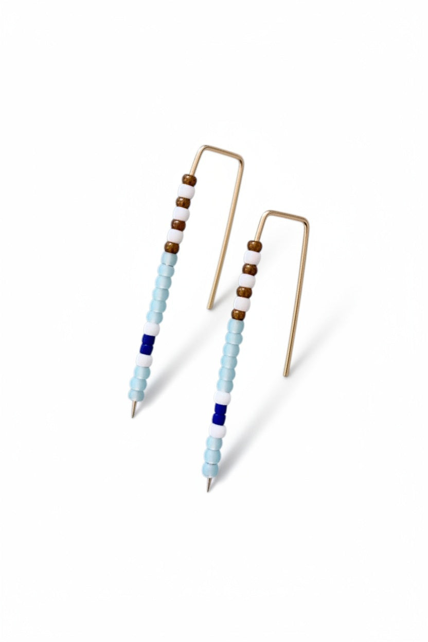 The ALICE RISE - BIG QUILL EARRINGS IN SKY BLUE feature gold wire and vertical rows of small beads in light blue, white, brown, and dark blue. Handmade by ALICE RISE, these earrings are perfect for sensitive skin and displayed on a textured light gray surface.