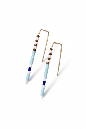 The ALICE RISE - BIG QUILL EARRINGS IN SKY BLUE feature gold wire and vertical rows of small beads in light blue, white, brown, and dark blue. Handmade by ALICE RISE, these earrings are perfect for sensitive skin and displayed on a textured light gray surface.
