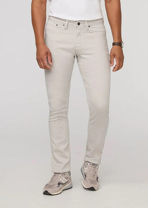 A person stands facing forward on a plain light background, wearing DUER NO SWEAT ATHLETIC TAPER PANTS IN MIST and matching light gray sneakers, feet slightly apart.