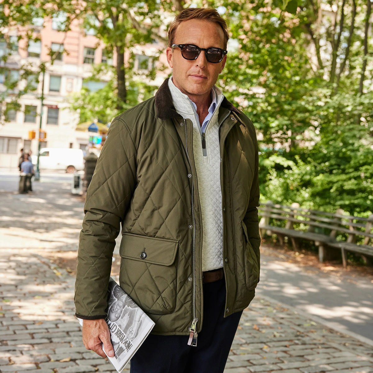 A man with light brown hair smiles while standing outside in a park. He wears the COLLARS AND CO - MIDDLEBURG QUILTED FIELD JACKET by COLLARS AND CO, featuring a corduroy collar, layered over a light blue shirt and tan pants for lightweight warmth on a cool day. Trees and a stone wall are visible in the background.