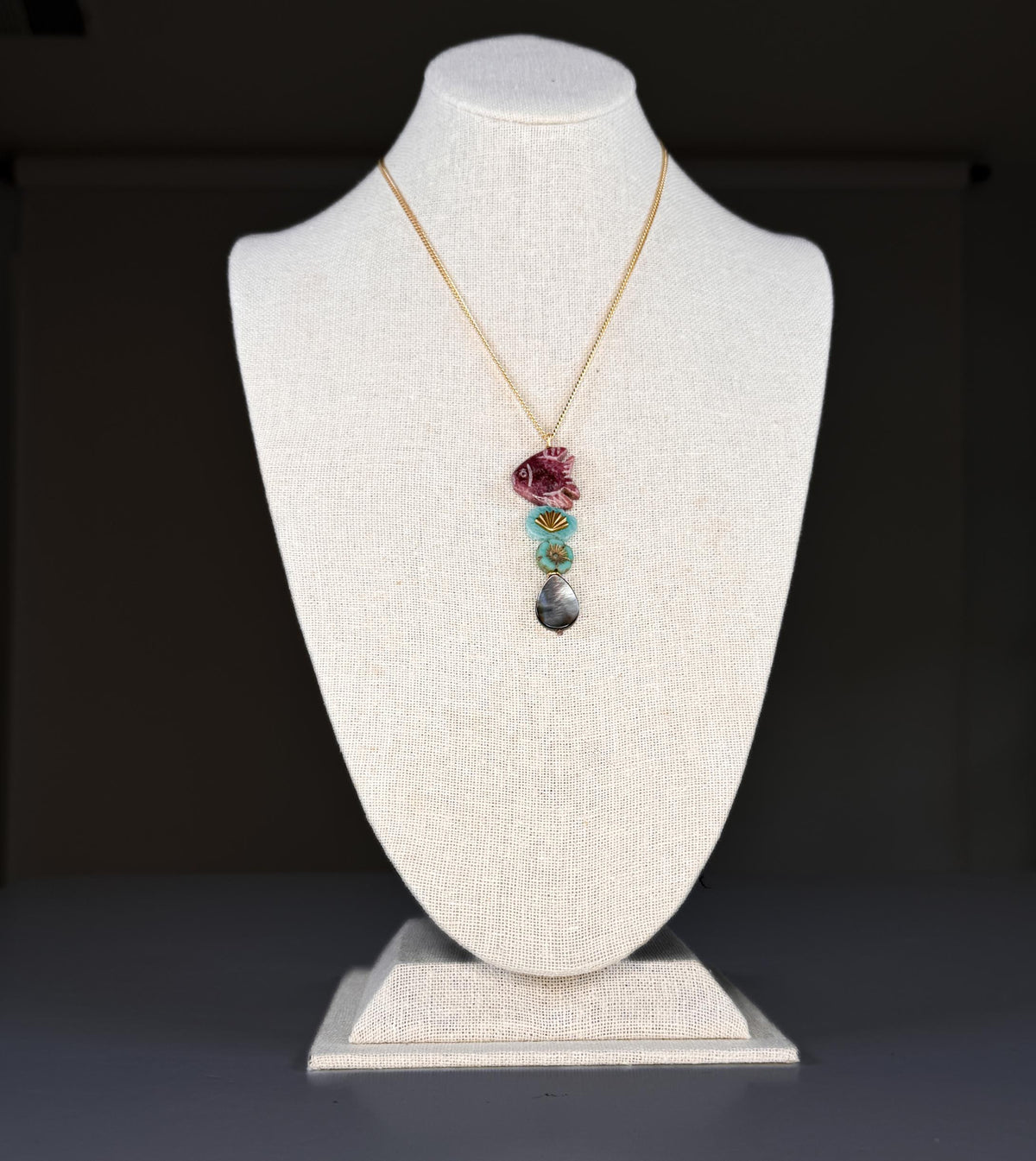 The SANDRINE FRANCE STUDIO - COMPANION LEO NECKLACE features a rhodonite fish pendant, two green Peruvian aventurine beads, and a teardrop-shaped gray stone on a gold chain—an elegant handmade piece from SANDRINE FRANCE STUDIO.