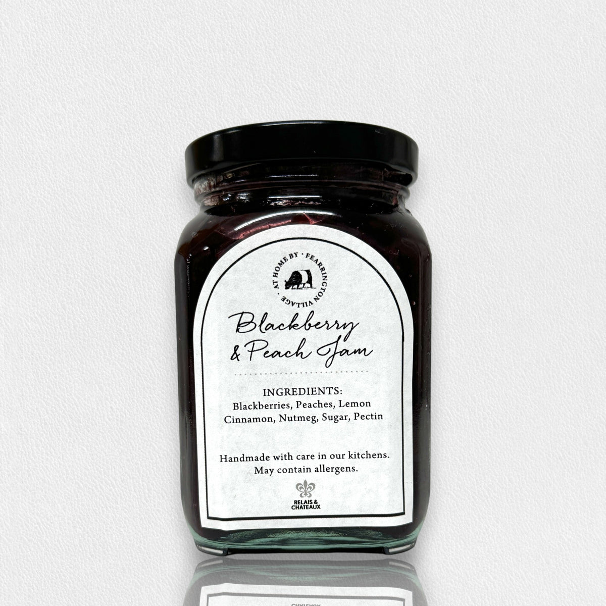A glass jar of FEARRINGTON SIGNATURE COLLECTION - FEARRINGTON HOUSEMADE BLACKBERRY PEACH JAM with a black lid and a white label. The label lists ingredients: blackberries, peaches, lemon, cinnamon, nutmeg, sugar, pectin. It also mentions: "Handmade with care in our kitchens by Fearrington chefs." Perfect as a hostess gift or for breakfast jams. May contain allergens.