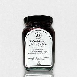 A glass jar of FEARRINGTON SIGNATURE COLLECTION - FEARRINGTON HOUSEMADE BLACKBERRY PEACH JAM with a black lid and a white label. The label lists ingredients: blackberries, peaches, lemon, cinnamon, nutmeg, sugar, pectin. It also mentions: "Handmade with care in our kitchens by Fearrington chefs." Perfect as a hostess gift or for breakfast jams. May contain allergens.