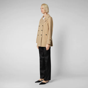 A person with short blond hair wears the SAVE THE DUCK - SOFI TRENCH JACKET by SAVE THE DUCK, a beige recycled polyester trench coat, over a white shirt with black pants and pointed shoes, standing against a light gray background.
