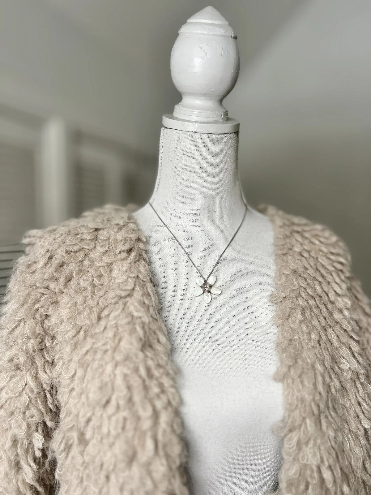 A white mannequin is dressed in a curly beige faux fur coat and adorned with the ANN LIGHTFOOT - SMALL MOTHER OF PEARL FACETED FLOWER NECKLACE by ANN LIGHTFOOT. The softly blurred background features light-colored walls and window blinds.