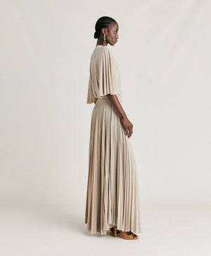 A person stands facing the camera, wearing the MOMONI - PARADISU DRESS, a light-colored, flowing pleated outfit with wide pants and kimono sleeves. Their hair is styled in neat braids, and they accessorize with drop earrings. The neutral background highlights the elegance of the MOMONI dress.