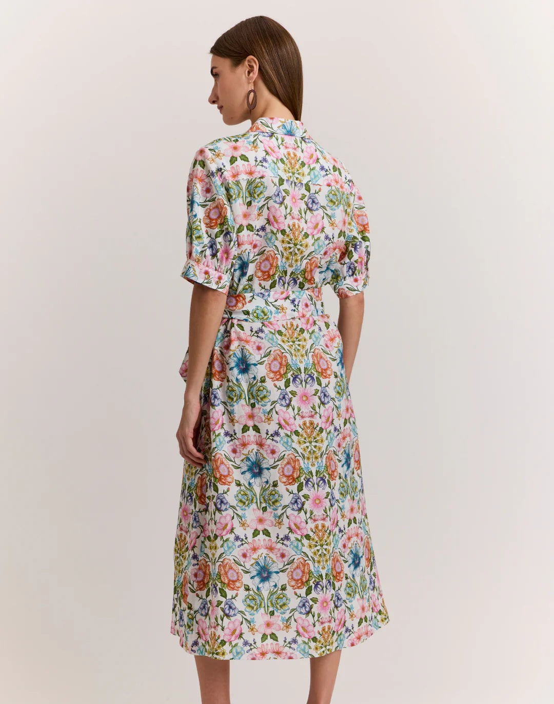A woman with straight brown hair stands sideways, wearing the HINSON WU Luca Elbow Sleeve Linen Blend Dress in Botanique, a light floral midi dress with short sleeves and a waist tie, set against a plain light background.