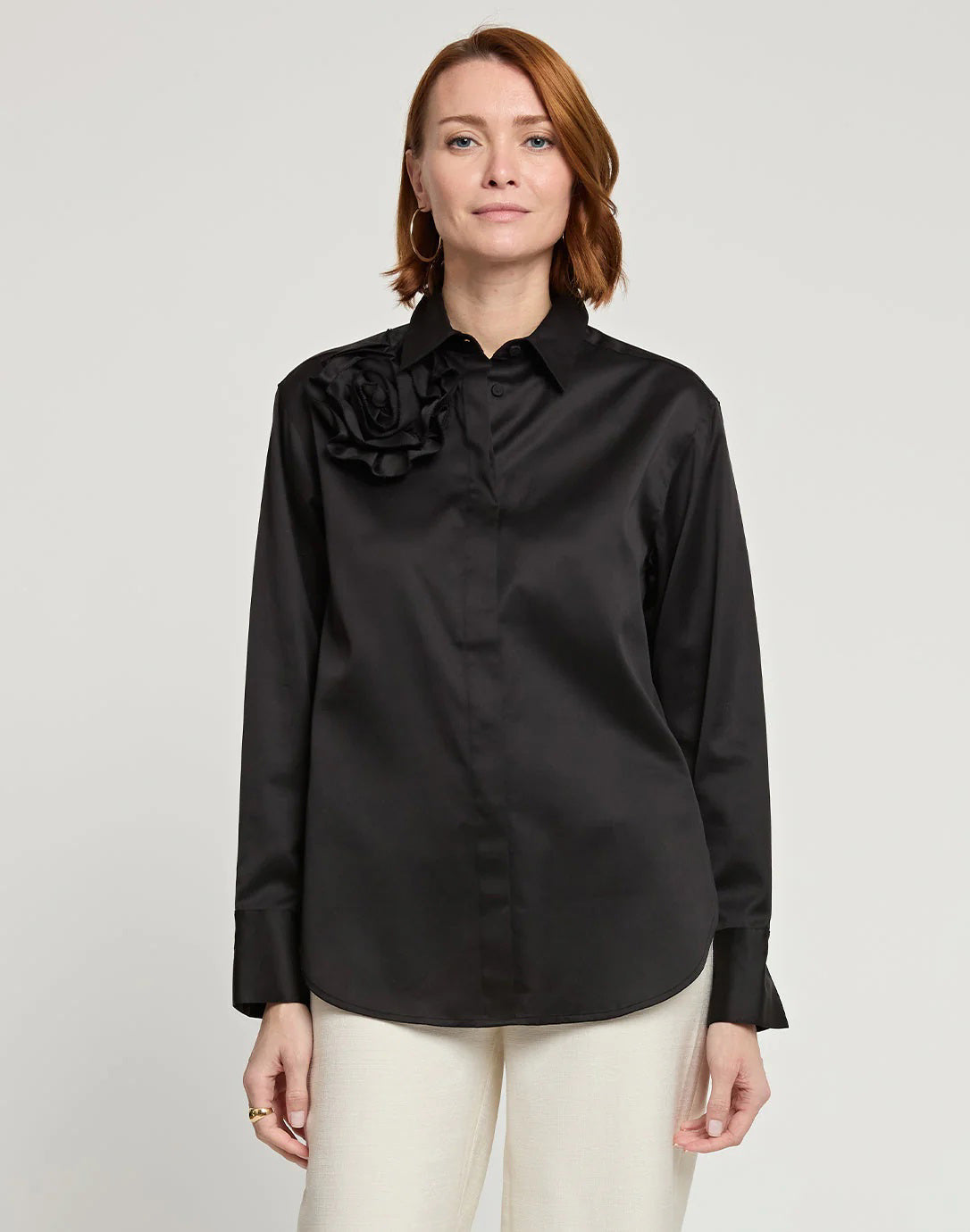 A person with short, brown hair is standing and facing away from the camera, wearing a HINSON WU - HALSEY LONG SLEEVE COTTON SATIN SHIRT WITH FLOWER. They have paired this black button-up shirt, made of a cotton and spandex blend, with light-colored pants. The background is plain and light-colored.