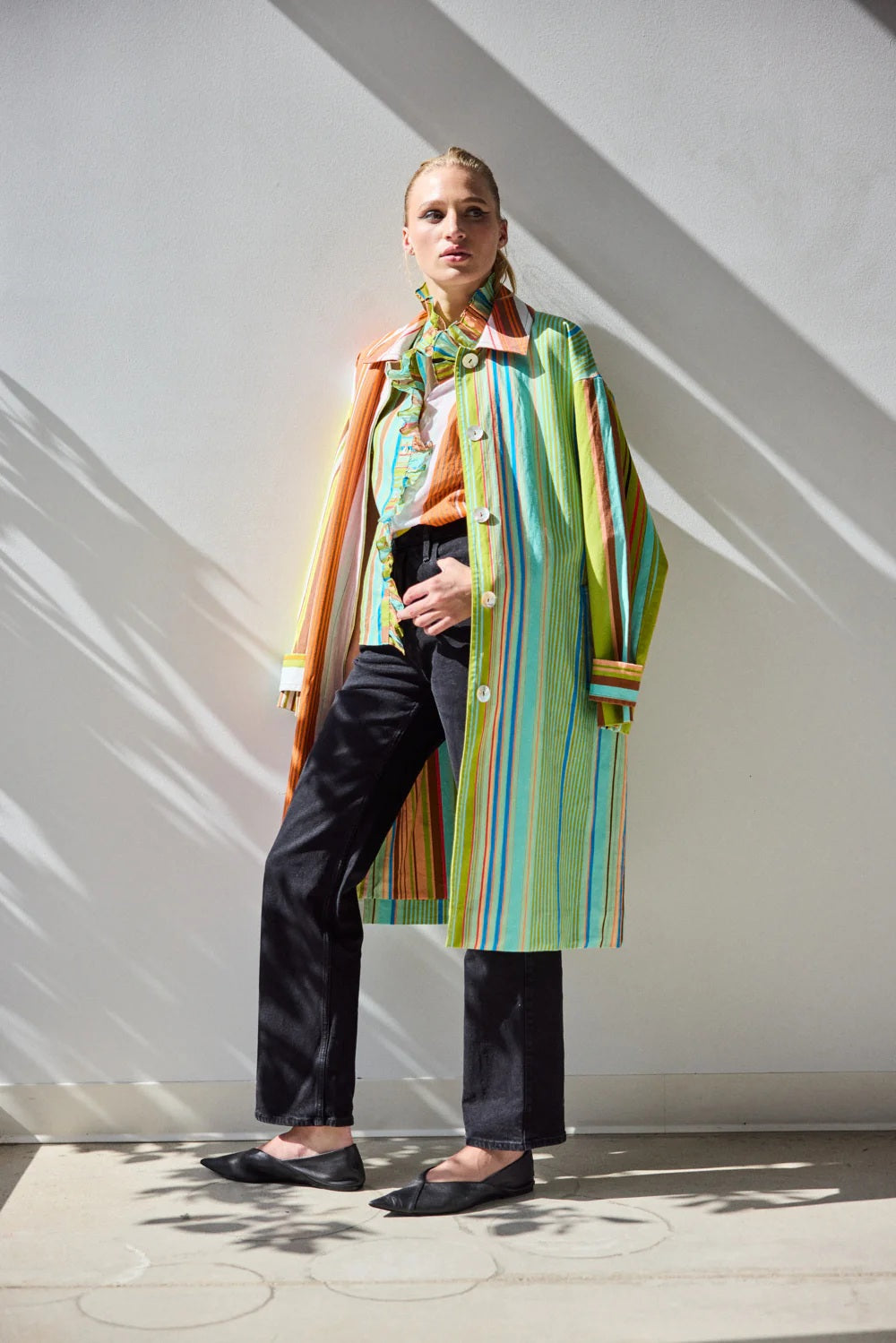 A person stands against a white wall wearing the HOPE FOR FLOWERS - COMBO STRIPES COAT by HOPE FOR FLOWERS, styled over a patterned shirt and black pants. Sunlight casts leaf-like shadows on the wall and floor behind them.