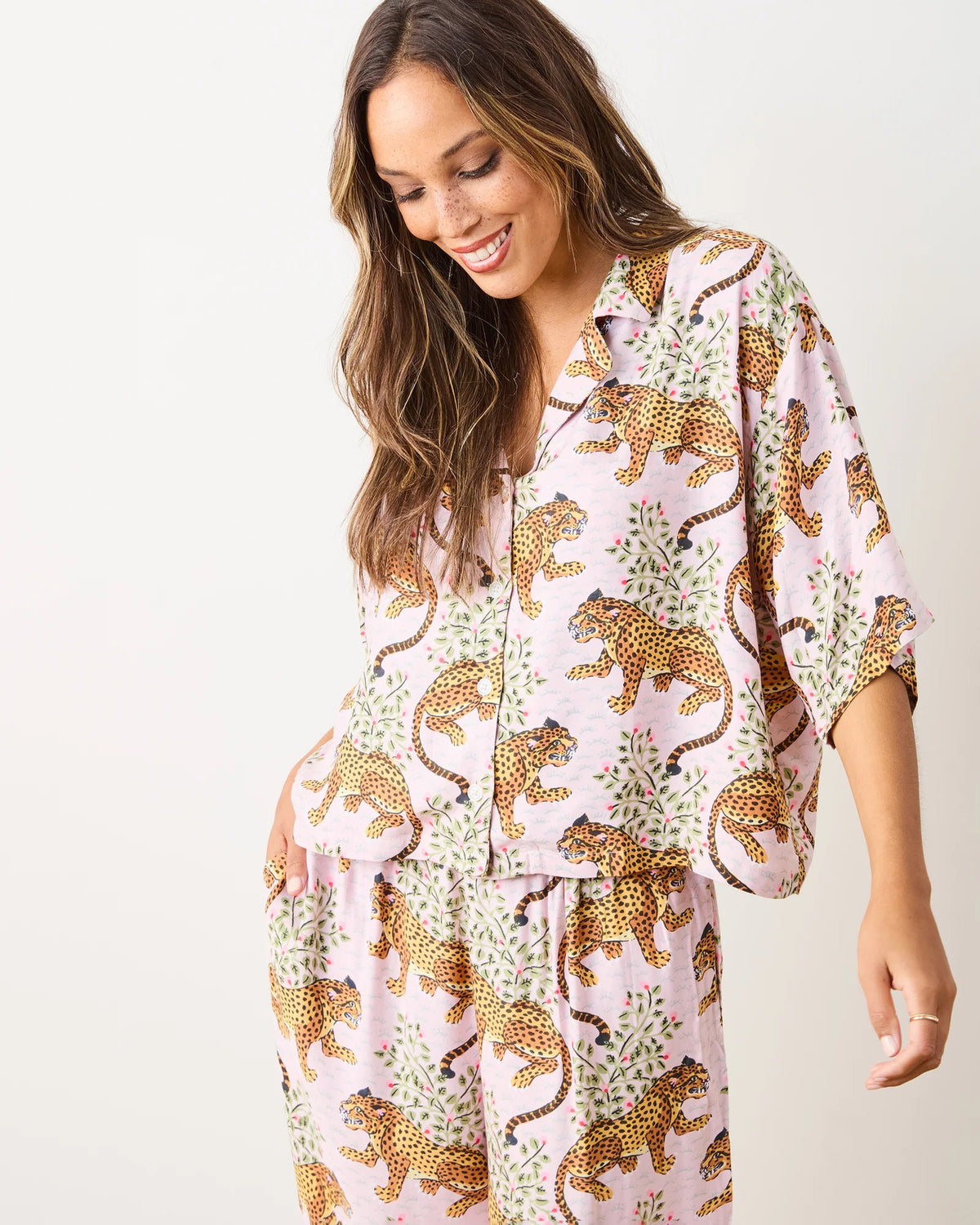 A person on a wooden floor wears the PRINTFRESH - Satin Bagheera Wildest Dream Pajama Set, made of eco-friendly viscose with a leopard print. The loose-fitting top and pants flow gently as they face away, presenting the back of their outfit against a plain, light-colored wall.