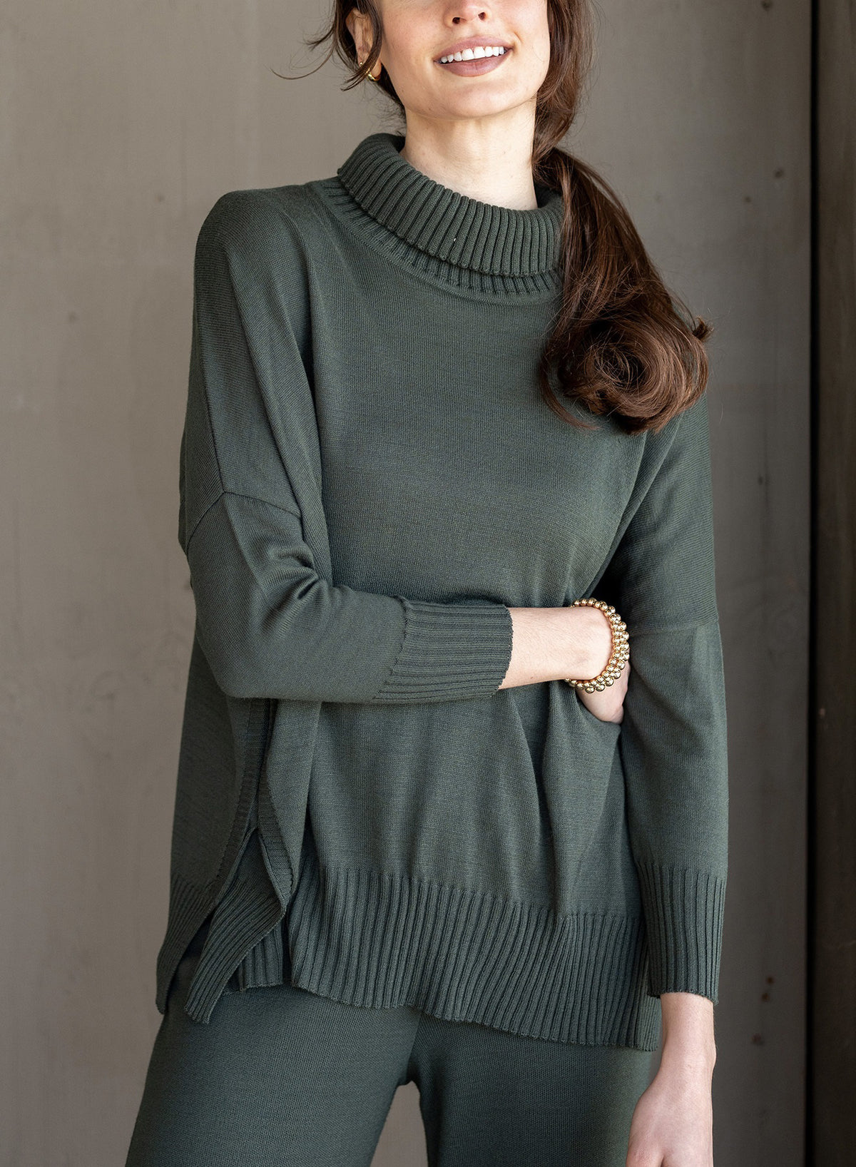 A woman with long brown hair wears the MERSEA Catalina Turtleneck Sweater in Deep Emerald with matching pants and a gold bracelet, standing against a neutral background with one hand on her hip and a slight smile.