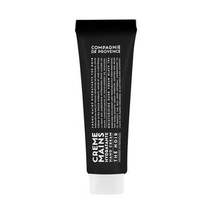 A black tube of COMPAGNIE DE PROVENCE - TRAVEL HAND CREAM BLACK TEA 1OZ from CIE LUXE BRANDS stands upright on a light, cylindrical stone pedestal against a neutral beige background with soft lighting.