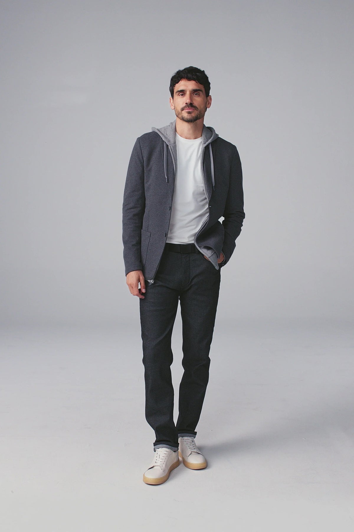 A man fastens the MACK WELDON Ace Blazer in Charcoal Heather over a gray hoodie, white tee, and dark jeans, paired with white sneakers with beige soles. He stands against a plain background, looking slightly to the side.