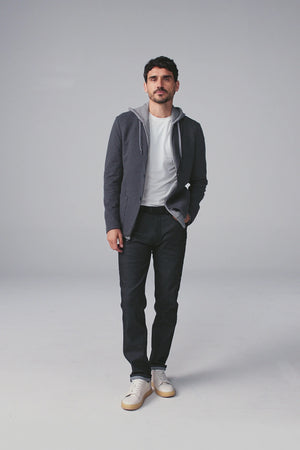 A man fastens the MACK WELDON Ace Blazer in Charcoal Heather over a gray hoodie, white tee, and dark jeans, paired with white sneakers with beige soles. He stands against a plain background, looking slightly to the side.