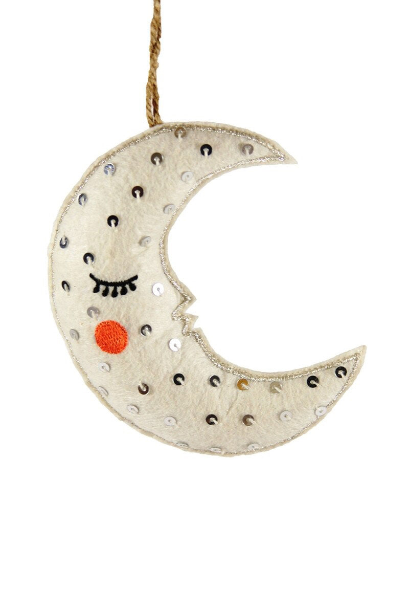 Introducing the HUSHED NIGHT MOON ORNAMENT from CODY FOSTER. This delightful felt crescent moon features gentle closed eyes and a cute red blush on its cheek. Crafted from soft cotton, it is beautifully embellished with shimmering silver sequins and elegantly hangs from a string.