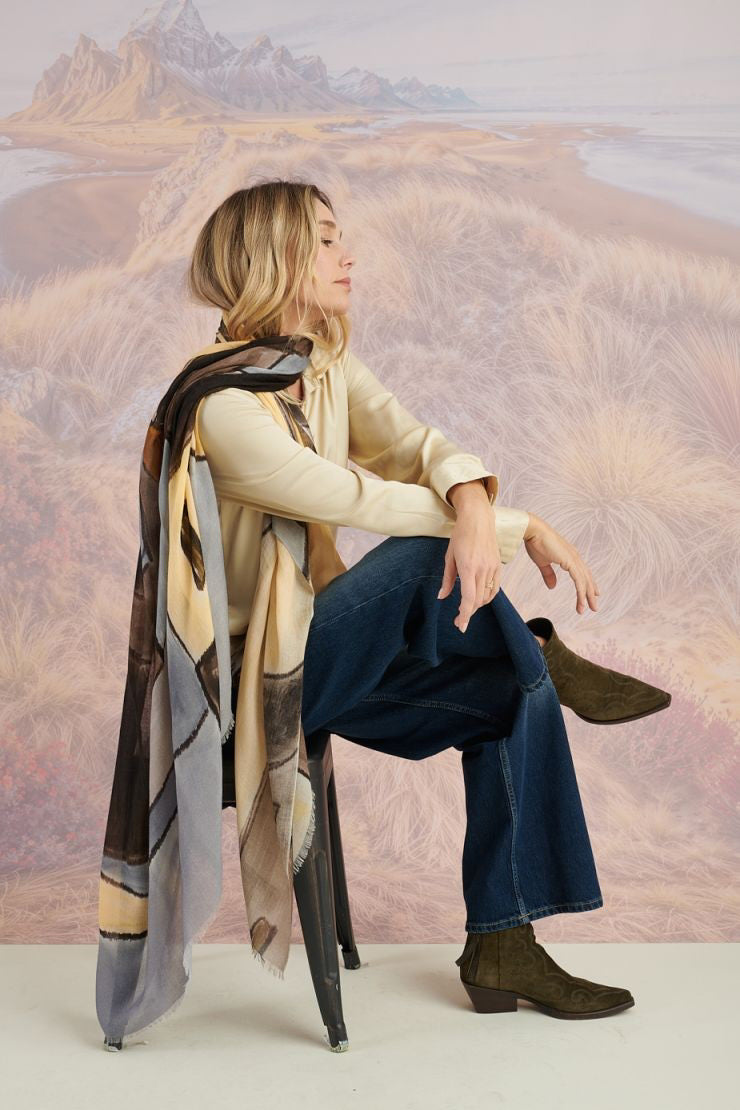 A woman in a black long-sleeve top and beige pants stands before a scenic backdrop, her look elevated by the STORIATIPIC ISCHIA Modal and Wool Scarf (100x200 cm) in vibrant orange and brown, draped elegantly around her neck.