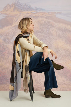 A woman in a black long-sleeve top and beige pants stands before a scenic backdrop, her look elevated by the STORIATIPIC ISCHIA Modal and Wool Scarf (100x200 cm) in vibrant orange and brown, draped elegantly around her neck.