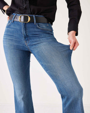 A woman stands against a plain white background wearing sunglasses, a black long-sleeve blouse, black heels, and holding a black handbag. She completes her look with the MERSEA Murphy Mini Boot Jean in Tide from MERSEA—blue, cropped organic cotton jeans designed for a gap-free waist.