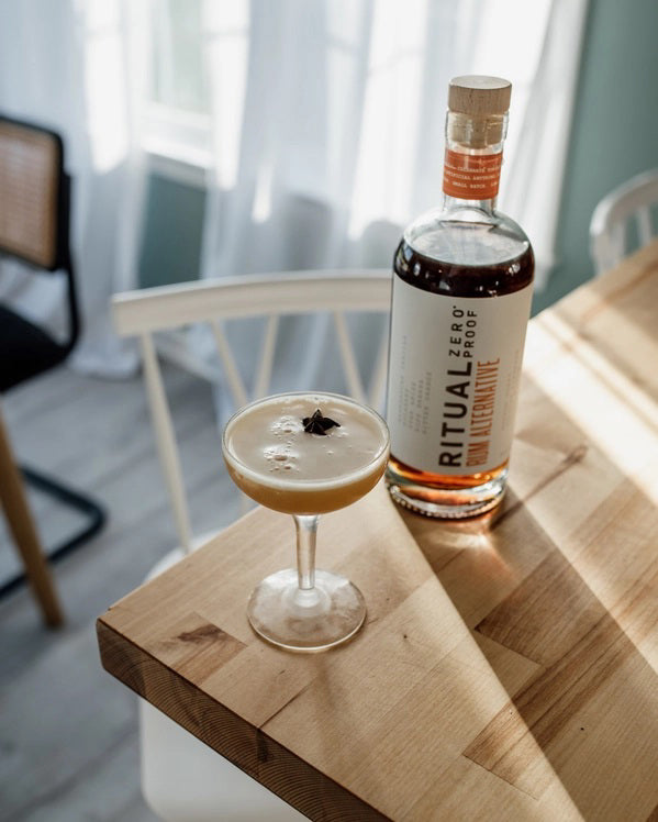 A bottle of RITUAL ZERO PROOF- RUM ALTERNATIVE by RITUAL ZERO PROOF, featuring a tan cap, amber liquid, and a white label with bold black and orange text describing this non-alcoholic rum alternative.