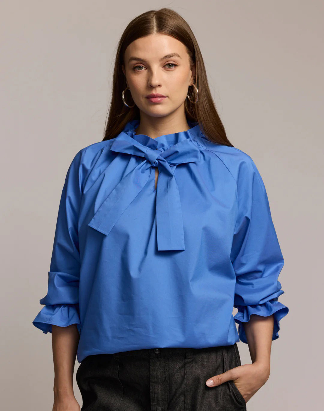 A woman with long brown hair models the HINSON WU ALINA LONG SLEEVE SHIRT, a bright blue blouse with a bow neck and ruffled sleeves, paired with dark pants. She stands against a light background, looking at the camera.