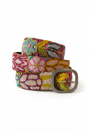 A person wearing a sleeveless beige knit dress and a JENNY KRAUSS - FOLKLORICA FLORAL EMBROIDERED BELT IN OLIVE with colorful floral patterns and a silver buckle.