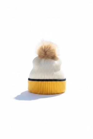 A woman, framed against a white background, wears the MERSEA - MONTAUK COLORBLOCK BEANIE IN CREME STRIPE—a cream knit hat with a yellow stripe and pom-pom—paired with a matching cream scarf and a chunky rib knit sweater accented by black stripes, showcasing the ultimate cold-weather accessory ensemble from MERSEA.