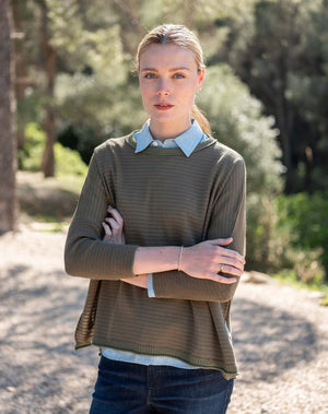 A woman with wavy brown hair stands barefoot on a sunlit floor near a window, smiling softly at the camera while wearing the MERSEA Catalina Crewneck Sweater in Jungle Stripe—an oversized green sweater by MERSEA layered over a blue shirt and paired with blue jeans.