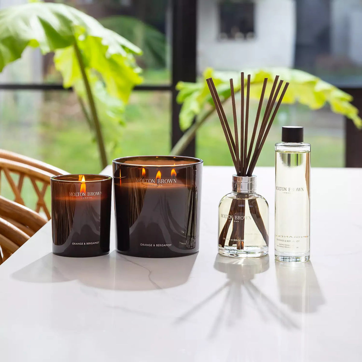 A Molton Brown - Orange and Bergamot Luxury Candle in a dark glass container. The label on the candle reads &quot;Molton Brown London&quot; at the top and &quot;Orange &amp; Bergamot&quot; at the bottom. Three wicks emit a citrus fragrance with notes of orange neroli.